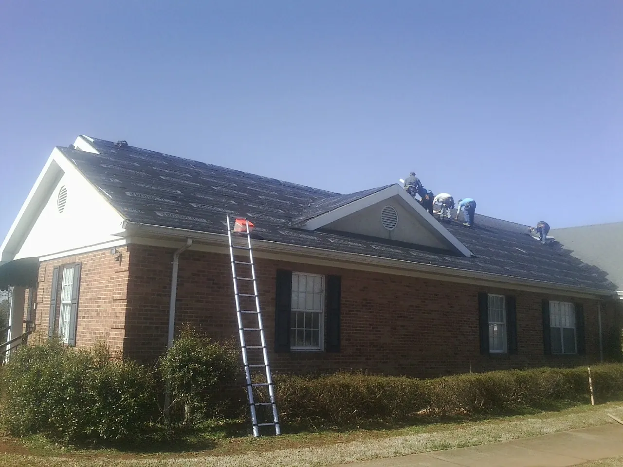 Crew tearing shingles off a brick ranch roof with a ladder set — Emergency Roof Repair work by our crew in Pipe Creek