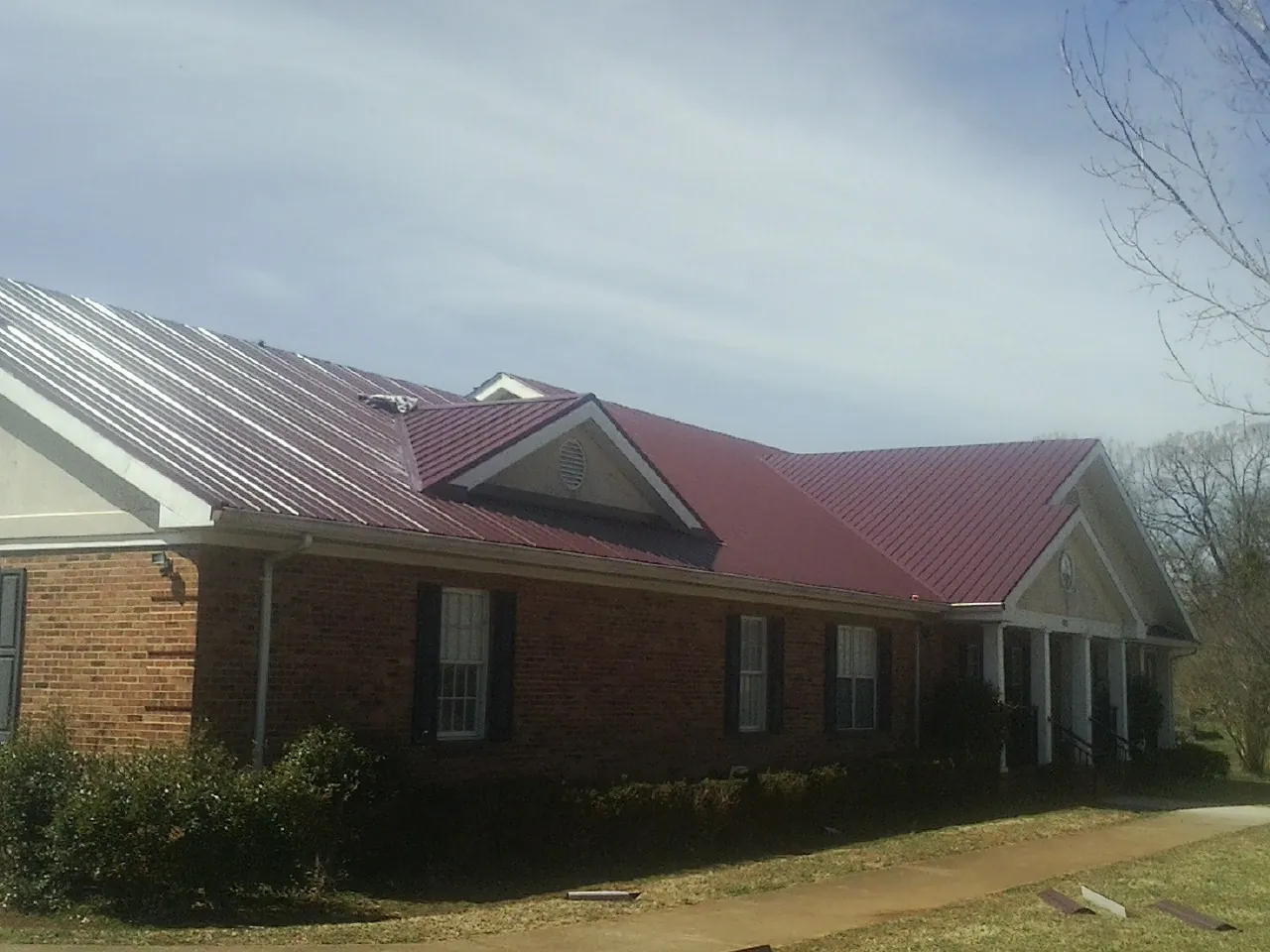 Red metal roof with gabled entries on a long brick building, from a Hail Damage Roof Repair job in Pipe Creek