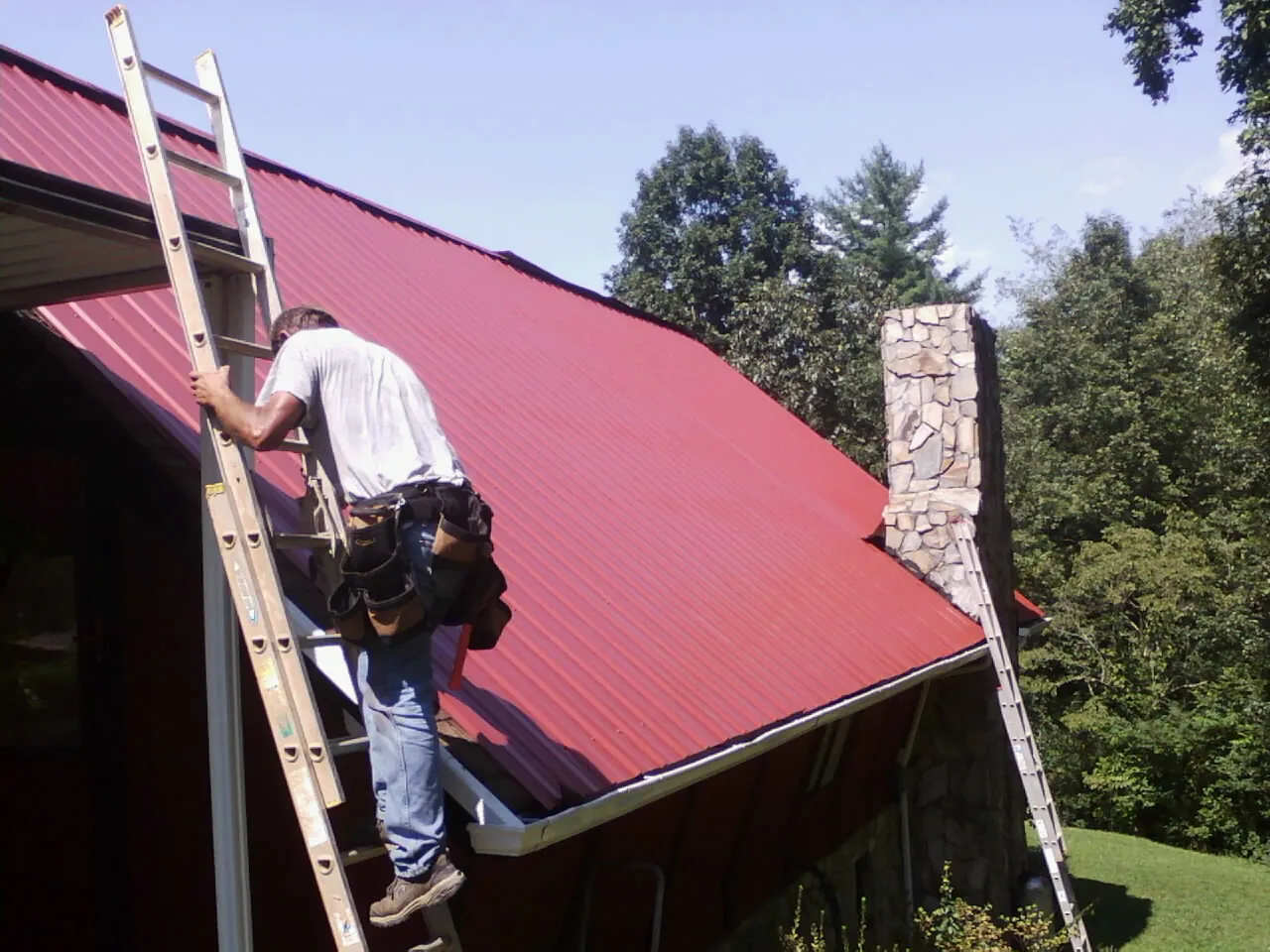 Roofer on a ladder beside a red metal roof and stone chimney — Soffit & Fascia Repair by Roofing Brothers in Pipe Creek